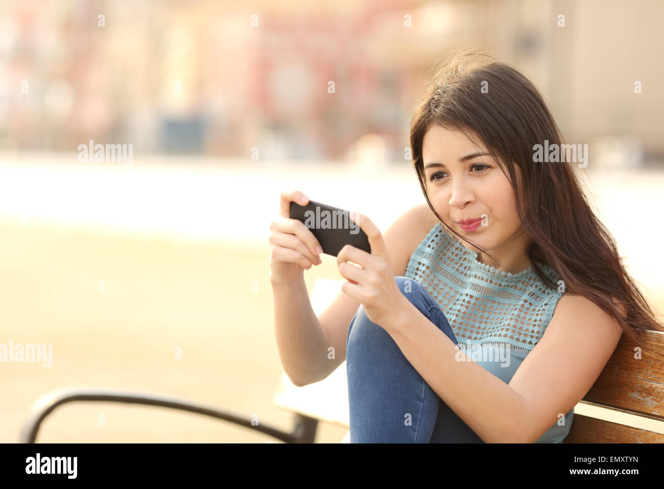 Funny teenager girl playing games on a smart phone sitting in a bench in a park Stock Photo
