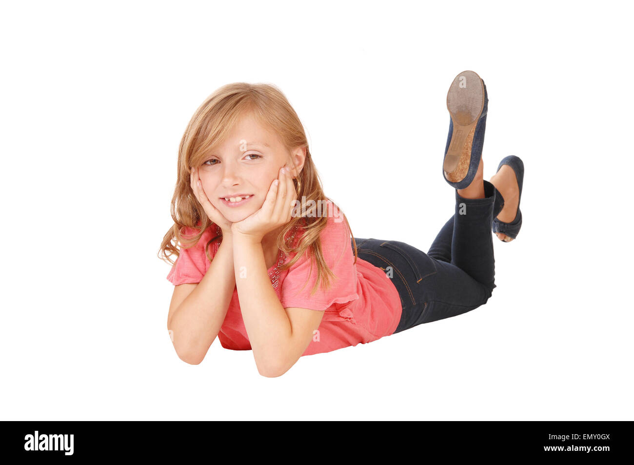A blond little girl in a pink sweater lying on the floor on her stomach, smiling, isolated on white background. Stock Photo