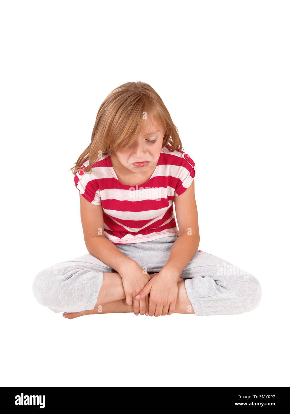 A little very unhappy girl sitting in her track pants and striped top on the floor and crying, isolated on white background. Stock Photo