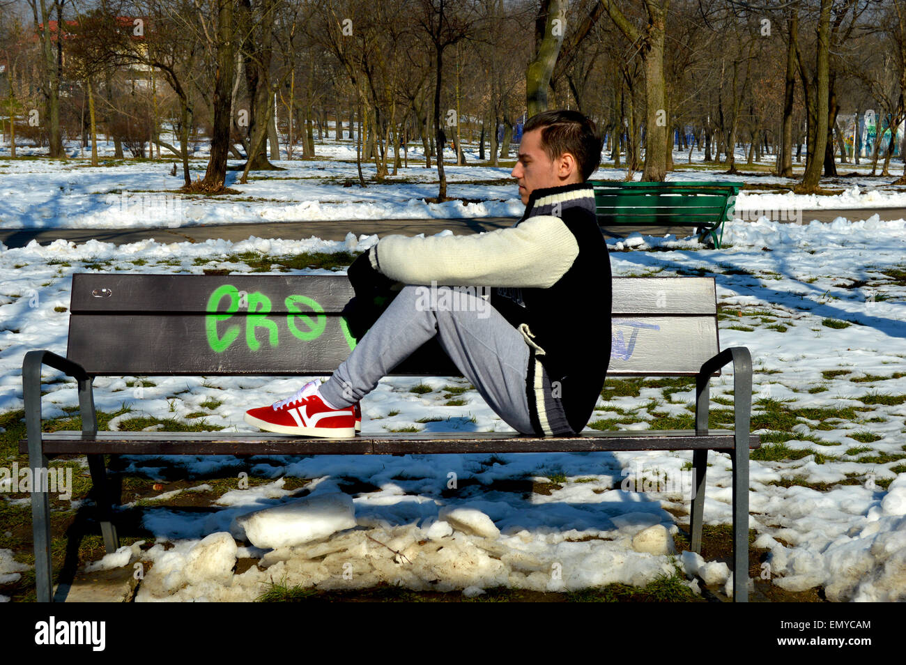 Man wearing hoodie, trainers and tracksuit pants sitting on park bench Stock Photo