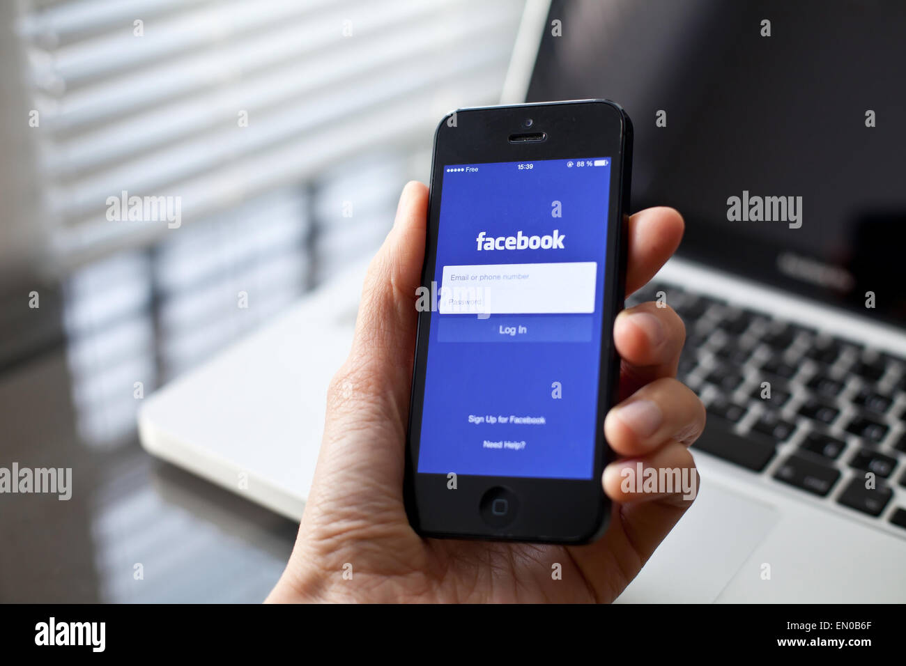 Hand holding Iphone with mobile application for Facebook on the screen, laptop on background Stock Photo