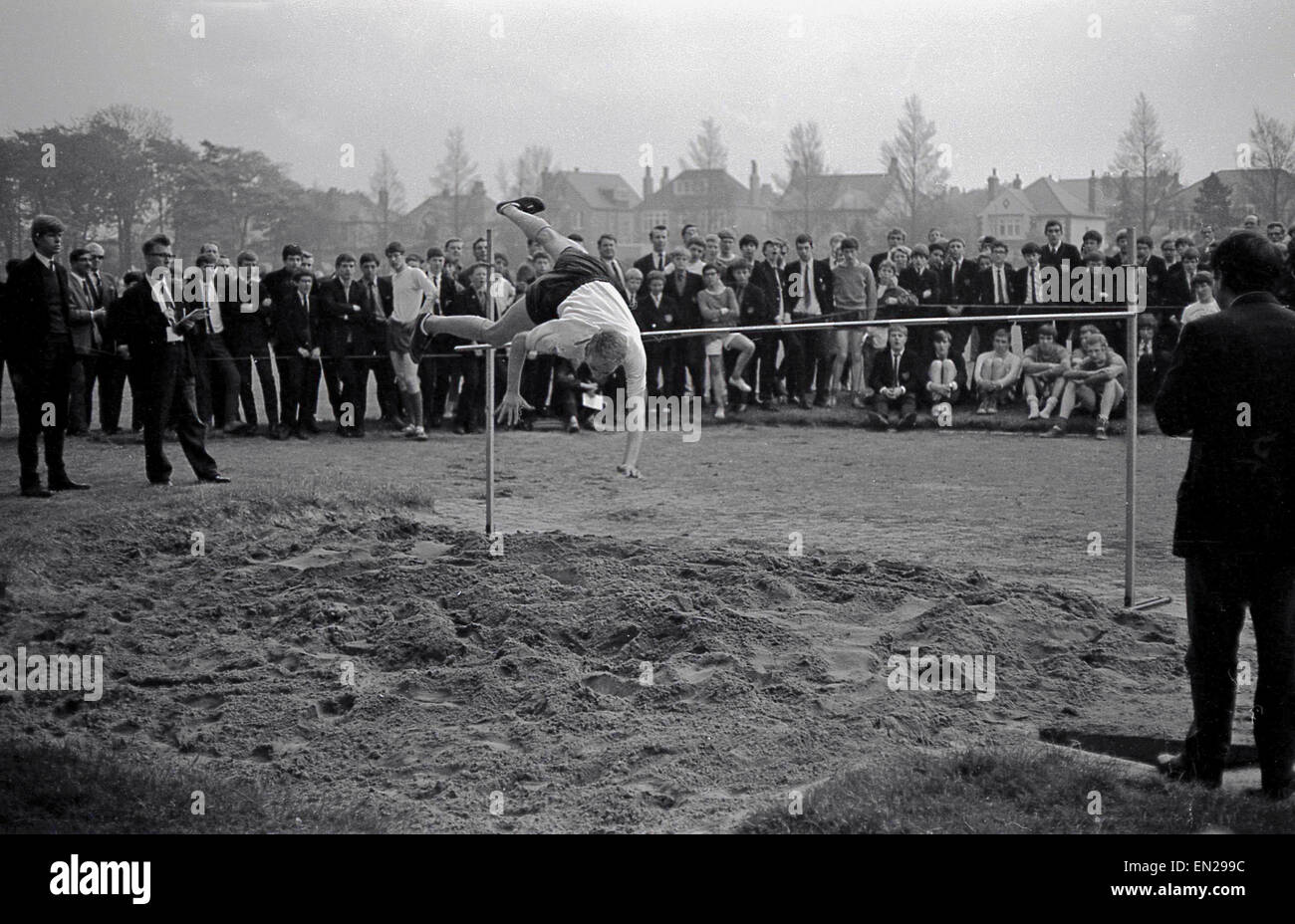 1960s historical picture, a school sports day sees a pupil attempting the high jump using the straddle or western roll technique, watched by fellow pupils. Stock Photo