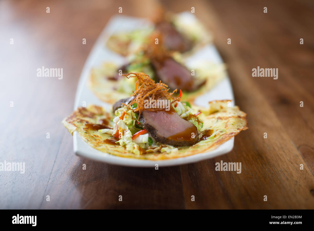 Three duck tacos on white plate with foreground in focus on wooden table Stock Photo