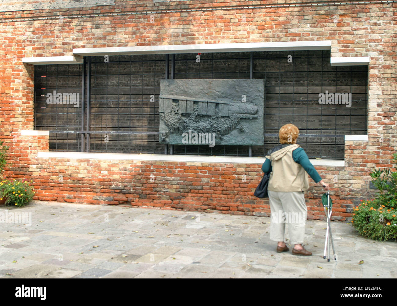 Venice, Province of Venice, ITALY. 7th Oct, 2004. A woman tourist stops to look at one of the Holocaust Memorials in the Campo del Nuovo Ghetto. In a cutout section of brick wall is a bronze bas-relief panel depicting The Last Train, with the names and ages of Venetian Jews killed by the Germans inside and behind it. Created by Arbit Blatas, a Lithuanian-born artist who lost his mother in the Holocaust, it was dedicated in 1993. Venice, a UNESCO World Heritage Site, is one of the most popular international tourist destinations. © Arnold Drapkin/ZUMA Wire/Alamy Live News Stock Photo