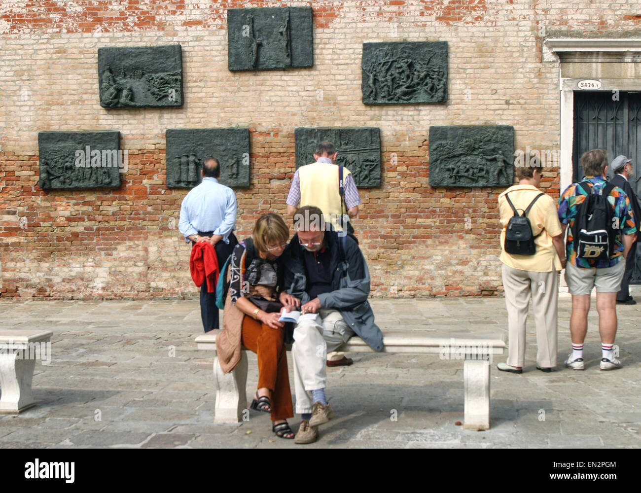 Venice, Province of Venice, ITALY. 7th Oct, 2004. A young couple on a bench read a guide book as other tourists view the seven-panel bronze bas-relief Holocaust memorial, on a wall of the Campo del Nuovo Ghetto by Arbit Blatas, a Lithuanian-born artist who lost his mother in the Holocaust. It commemorates the night of Dec. 5, 1943, when the first 200 of the city's Jews were rounded up and marched out for deportation and death. Venice, a UNESCO World Heritage Site, is one of the most popular international tourist destinations. © Arnold Drapkin/ZUMA Wire/Alamy Live News Stock Photo