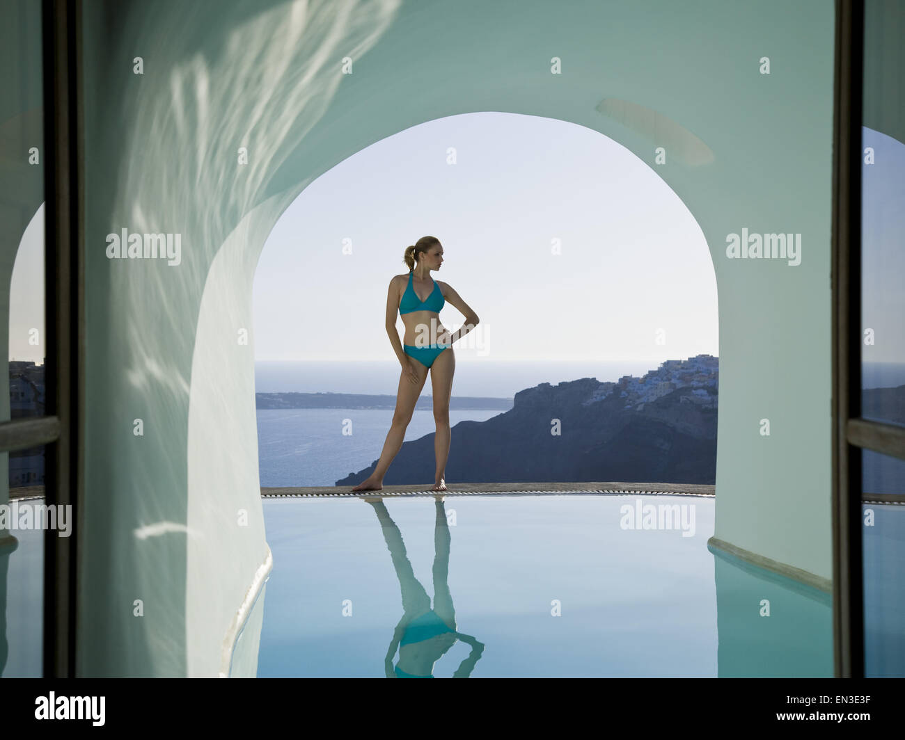 Woman in bikini standing at edge of infinity pool with arch and rock formation Stock Photo