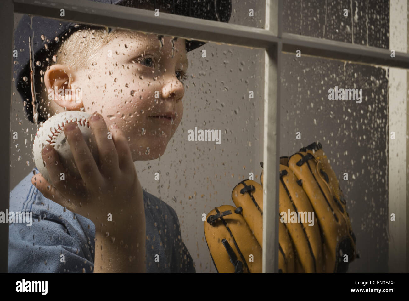 Young boy with baseball glove looking out window on rainy day Stock