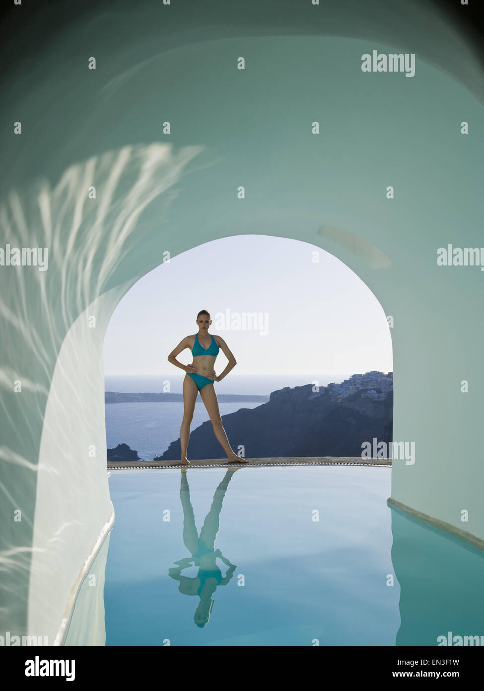 Woman in bikini standing at edge of infinity pool outdoors with arch and rock formation Stock Photo