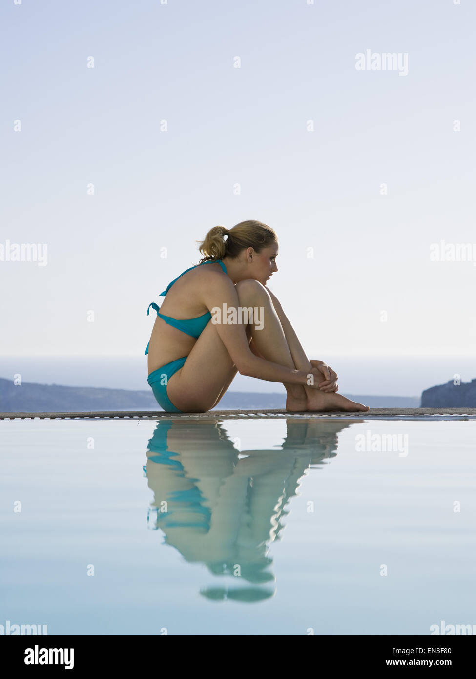Woman in swimsuit sitting at edge of infinity pool outdoors Stock Photo
