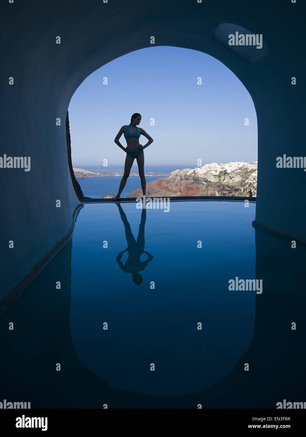 Silhouette of woman in bikini standing at edge of infinity pool with arch and rock formation Stock Photo