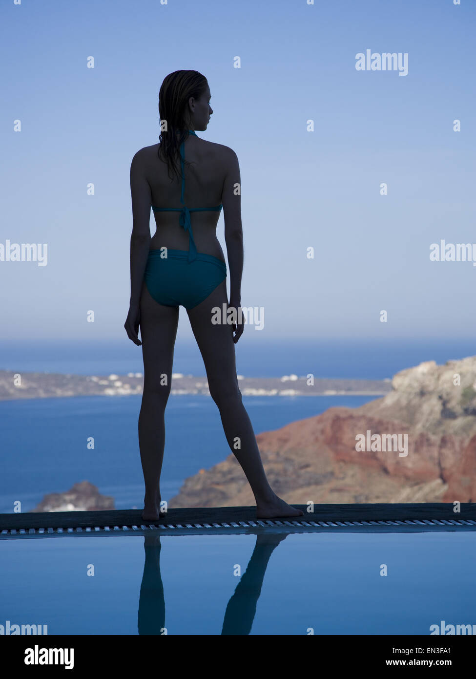 Rear view silhouette of woman in bikini standing at edge of infinity pool with rock formation Stock Photo