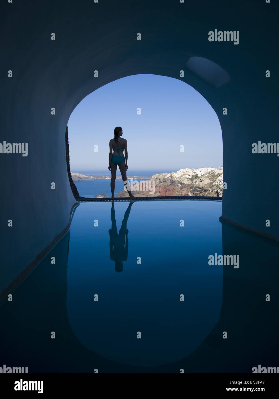 Silhouette rear view of woman in bikini standing at edge of infinity pool with arch and rock formation Stock Photo