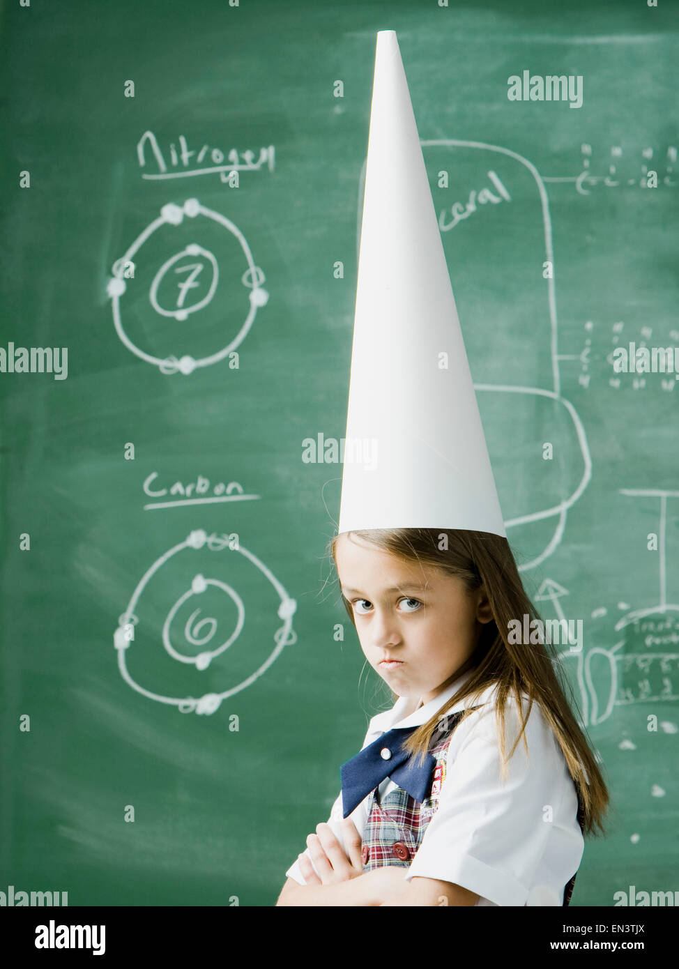 girl in a classroom standing in front of a chalkboard wearing a dunce