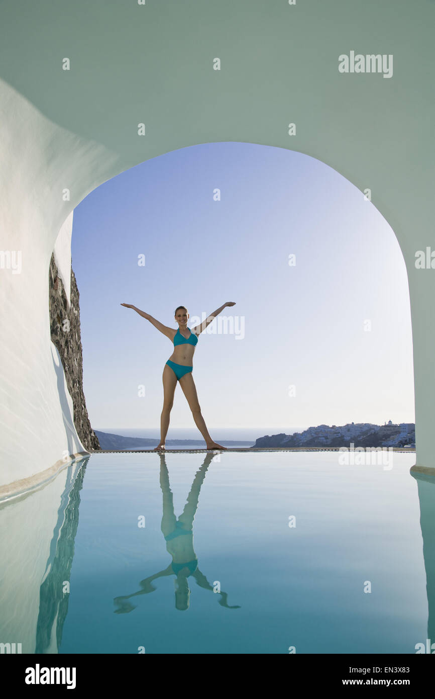 Woman in swimsuit standing at edge of infinity pool with arms up Stock Photo