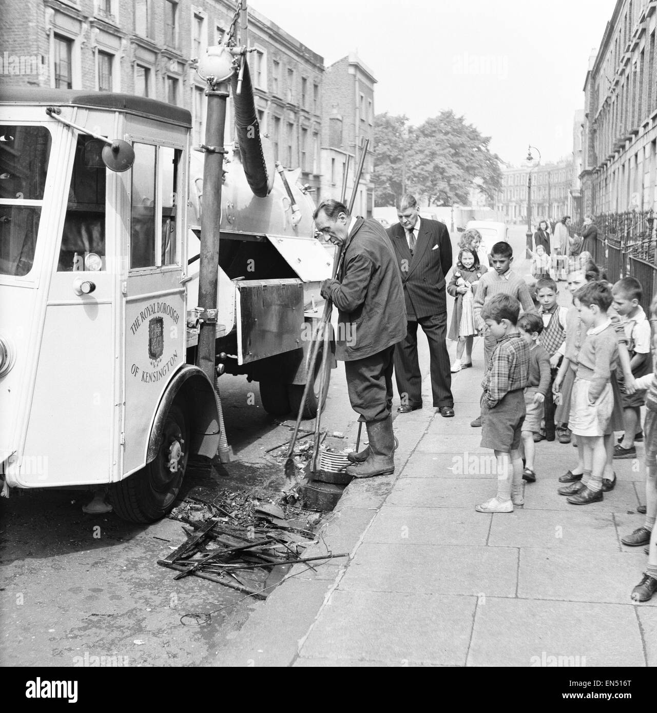 Two detectives surround by children watch as council workman searches a drain for the weapon that killed West Indian Kelso Cochrane 32. Cochrane was murdered by a group of teenagers in a street on the fringes of Notting Hill. 22nd May 1959 Stock Photo