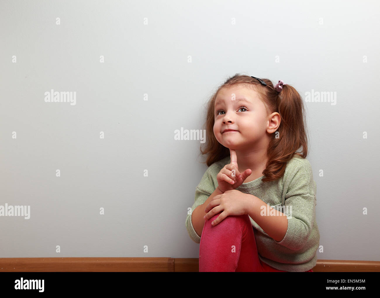 Fun thinking kid girl sitting in pink jeans and looking up on blue copy space background Stock Photo