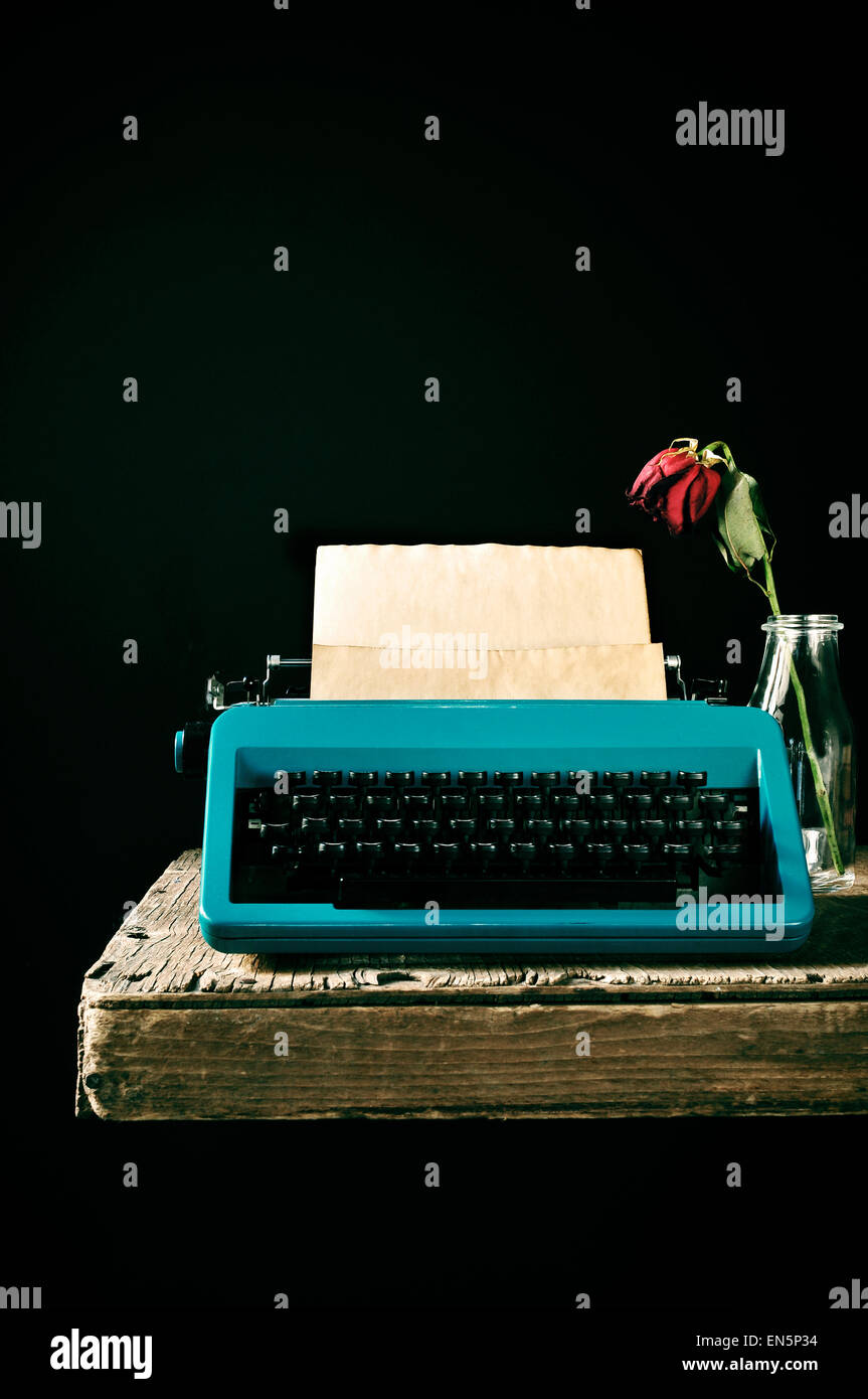 an old blue typewriter with a blank page in its roller and a wilted red rose in a glass vase on a rustic wooden table, against Stock Photo