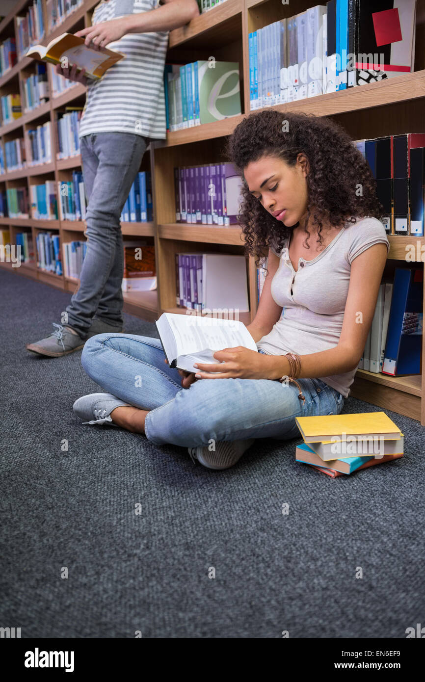 Pretty student sitting on floor reading book in library Stock Photo - Alamy