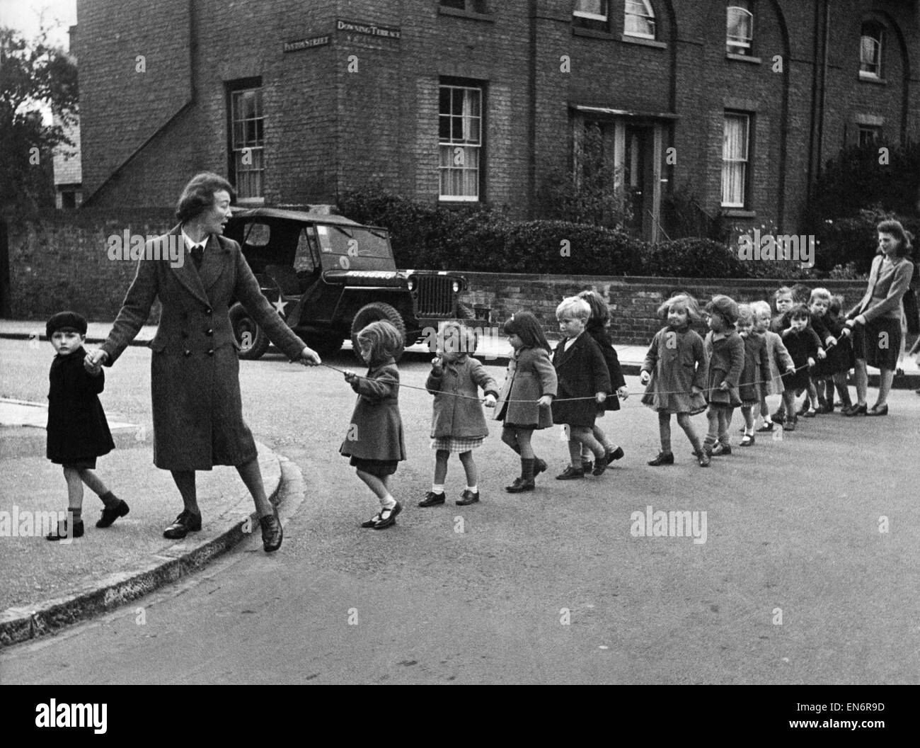 War workers' children set out for a morning stroll in Cambridge. They come form the Norwich St. Nursery. The rope serves two purposes; it keeps the toddlers together and by keeping them on the inside of the rope they cannot run into the road. October 1944 Stock Photo