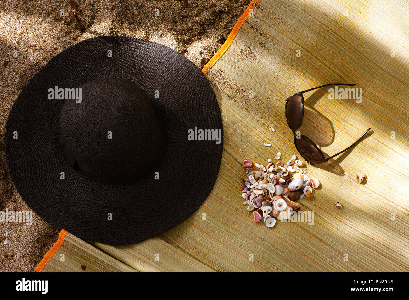 A natural fiber mat, sun hat, sun glasses and collection of shells at the beach, shot from above. Stock Photo