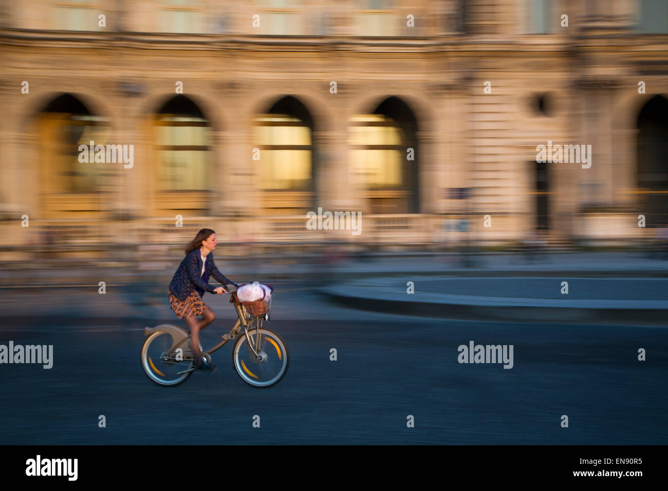 Panning photo of woman riding bicycle past Musee du Louvre at sunset, Paris, France Stock Photo