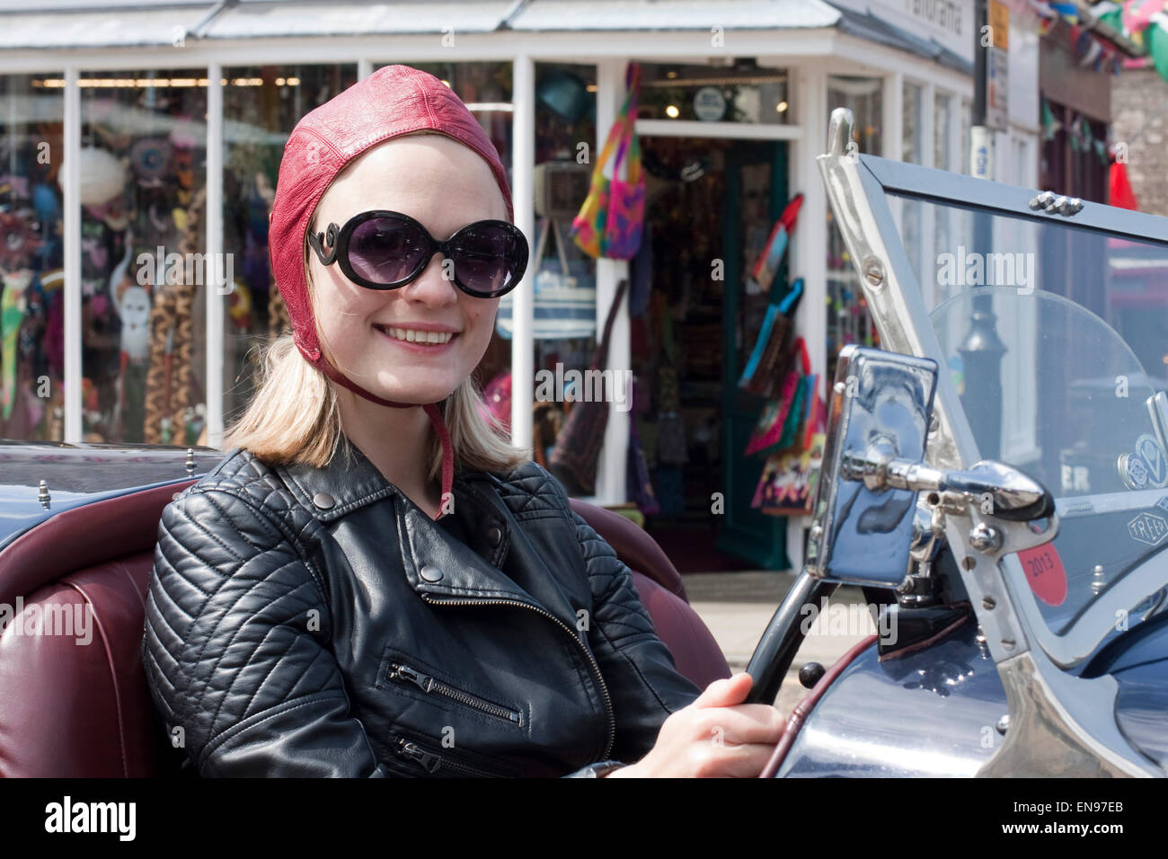 Young woman in black leather biker jacket and red flying helmet at the wheel of a open top vintage Riley Stock Photo