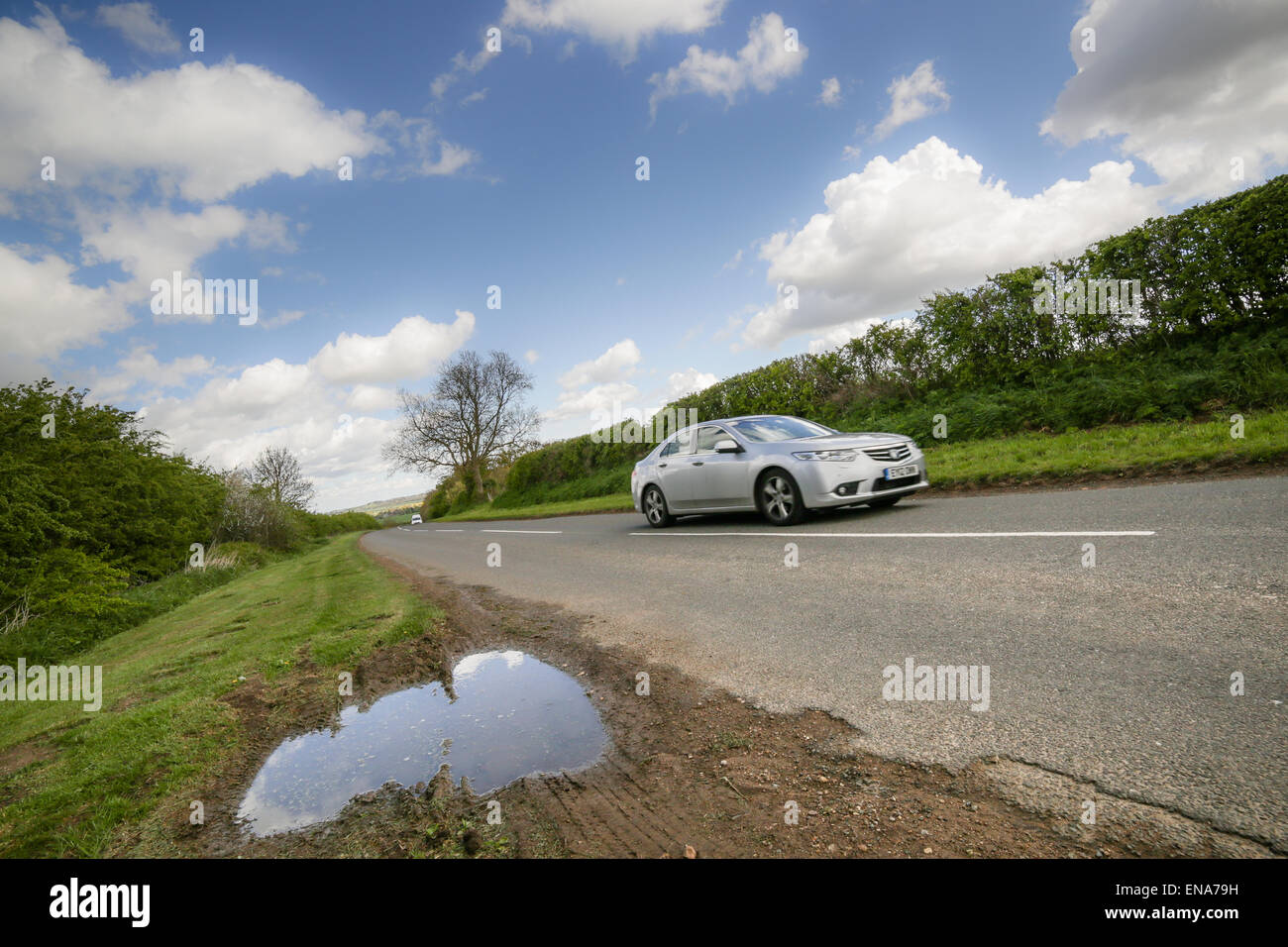 A silver saloon car rushes down a country road,past a puddle Stock Photo