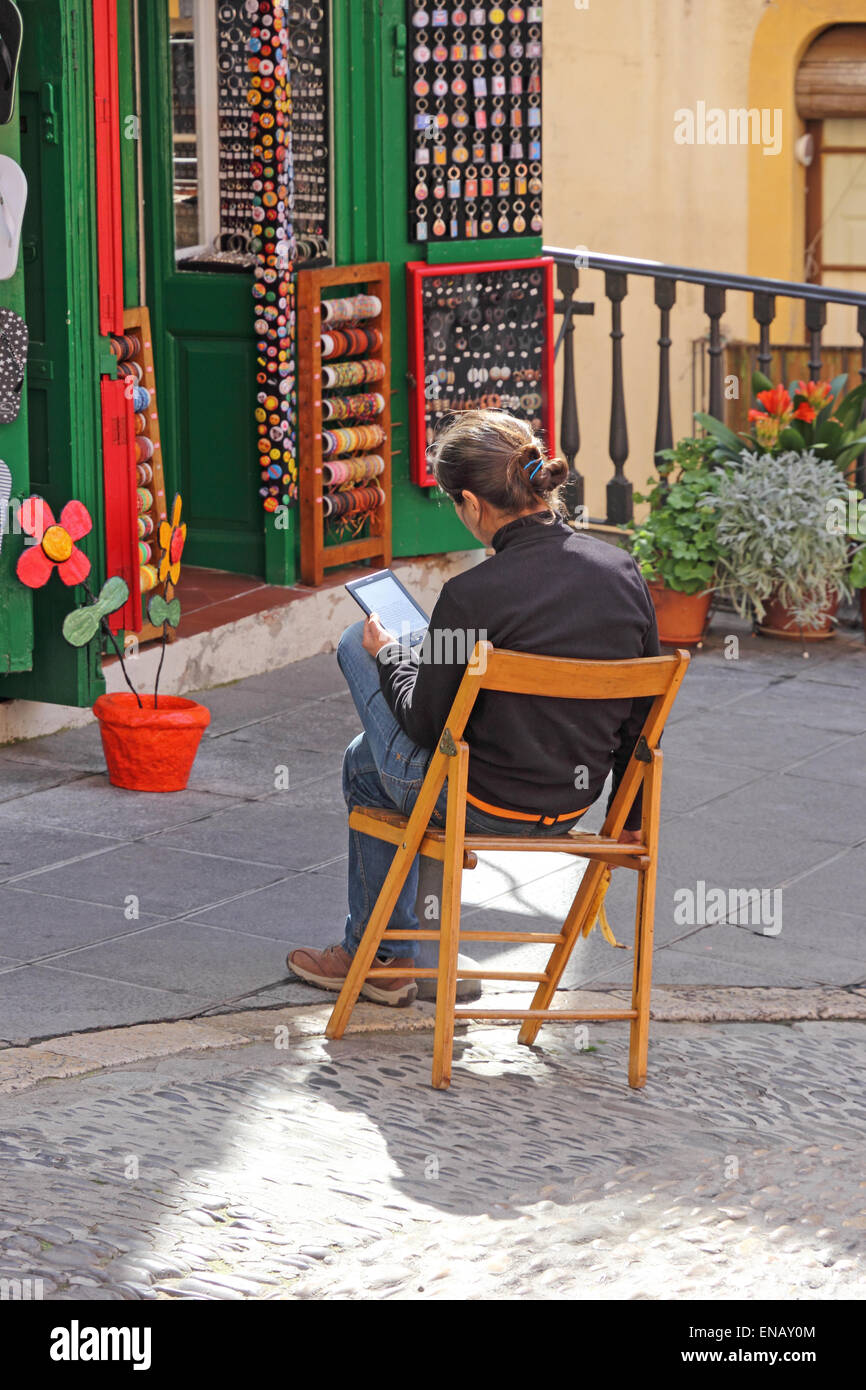 Young lady sat in sun outside souvenir shop, using tablet device, Tarragona, Spain Stock Photo