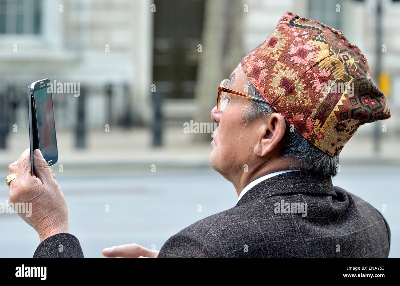 London, England, UK. Old Nepalese man (retired Gurkha) wearing a traditional Dhaka Topi (hat) taking a photo on his mobile phone Stock Photo