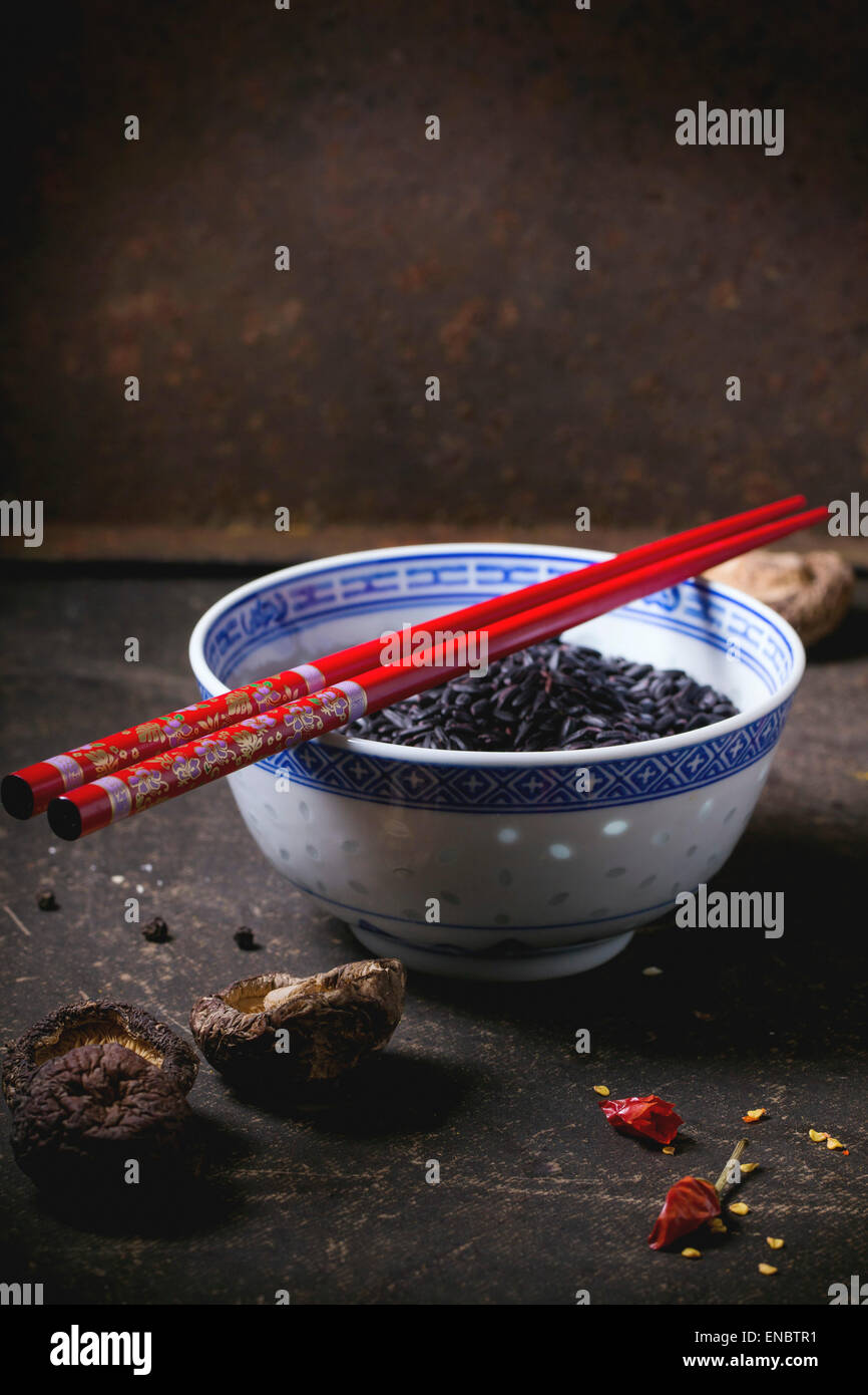 Porcelain bowl of uncooked black rice, served with dry shiitake mushrooms and red chopsticks over dark table. Stock Photo