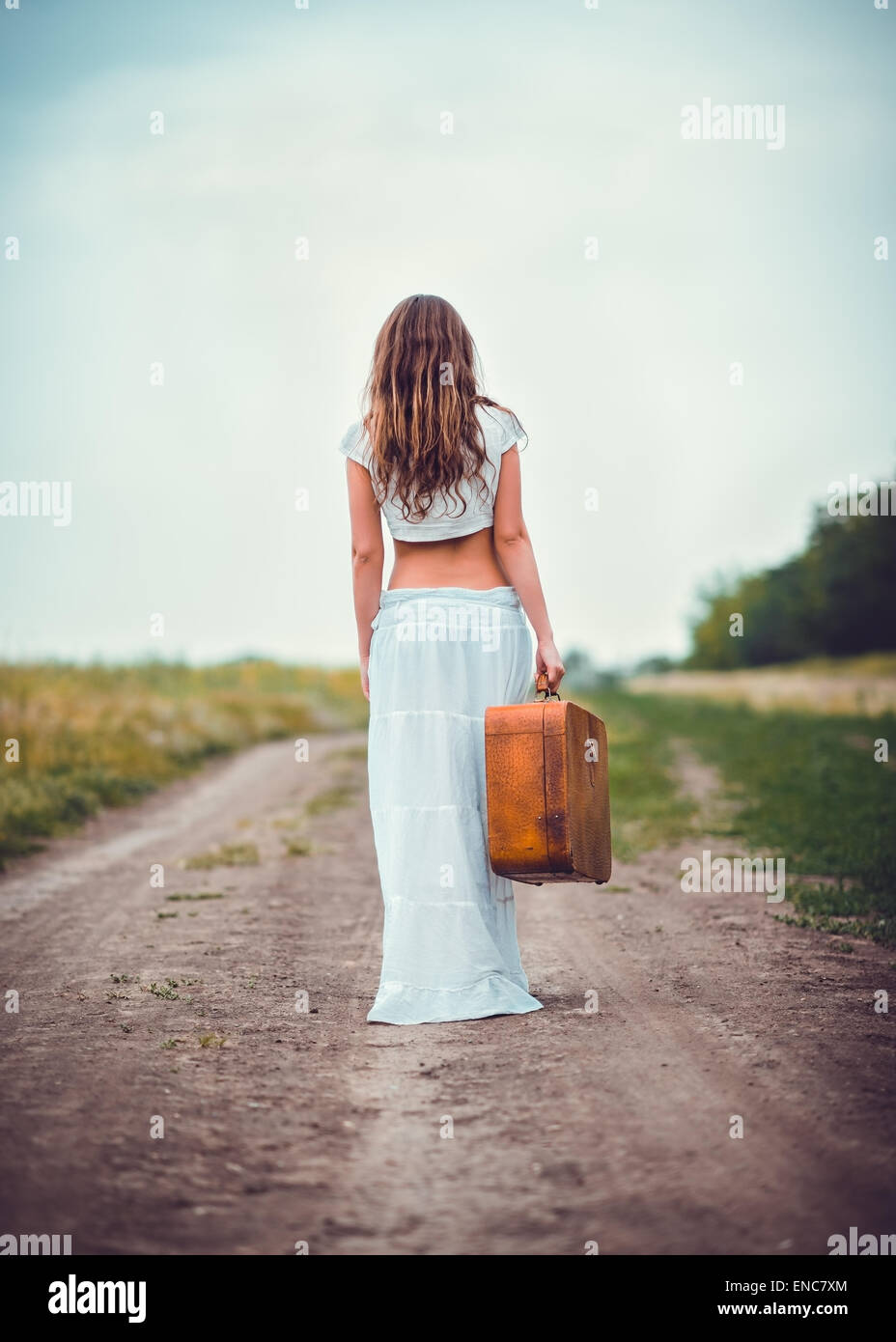 Young woman with suitcase in hand going away by a field road Stock Photo