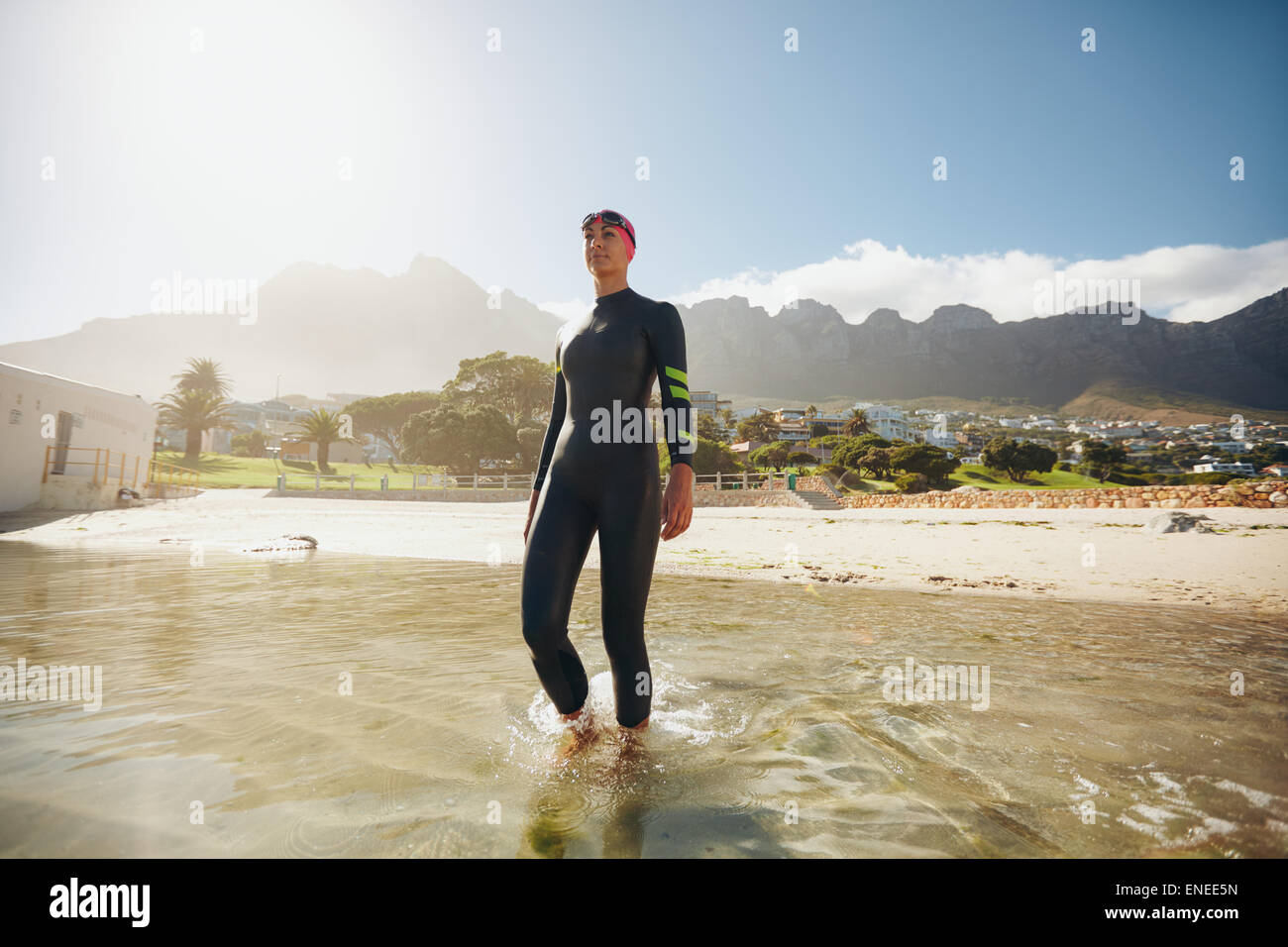 Image of fit young female triathlete walking into the sea wearing wetsuit. Triathlete in training at the beach. Stock Photo