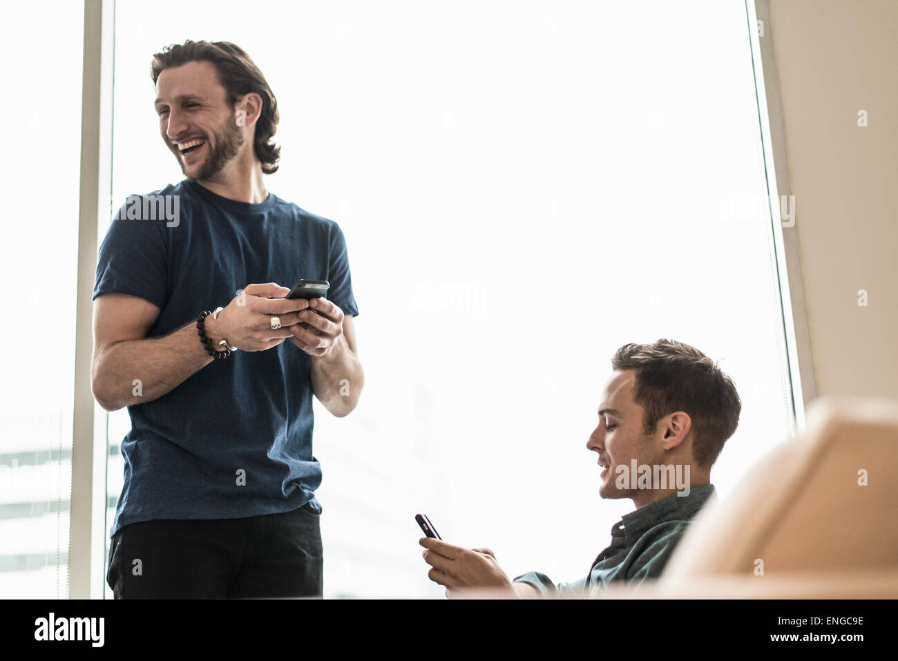 Two men in an office, checking their smart phones. One looking away laughing. Stock Photo