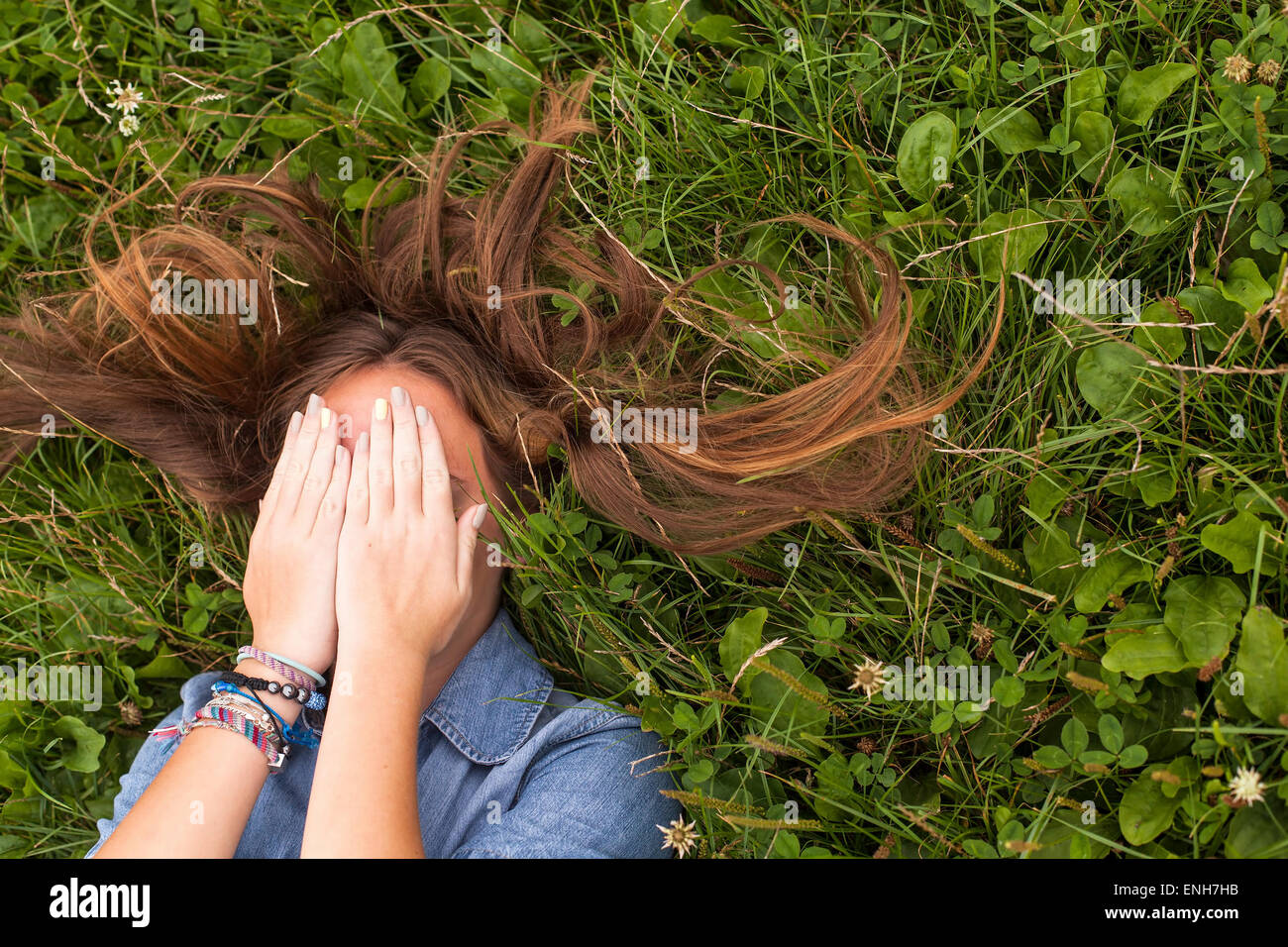 Young cute girl with tousled long hair lying on the green grass covering his face with his hands. Stock Photo