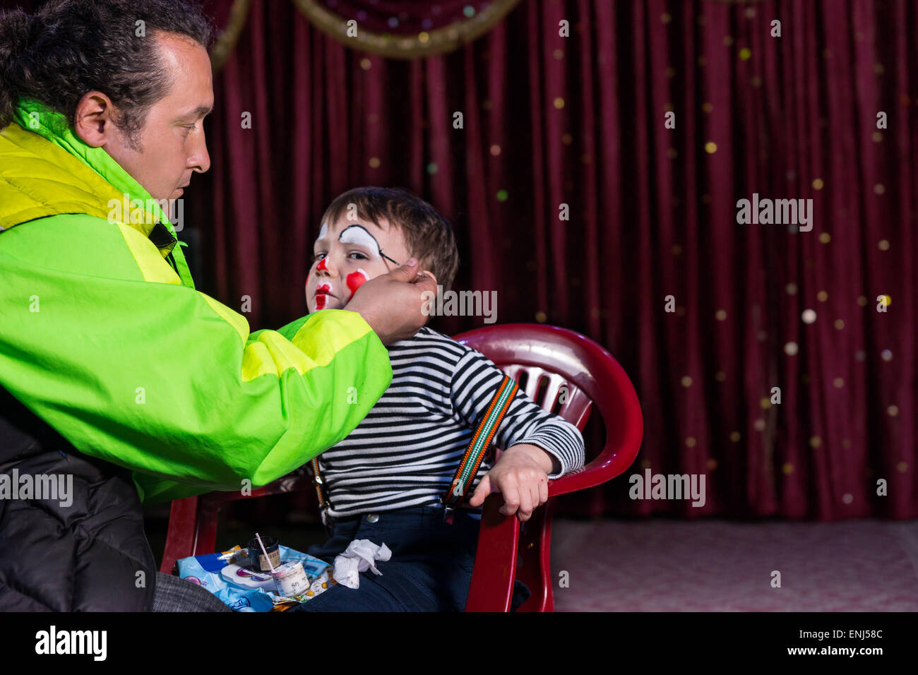 Boy Sitting in Chair on Empty Stage Having Clown Make Up Applied to Face by Man, Boy is Looking at Camera Out of Corner of Eye Stock Photo