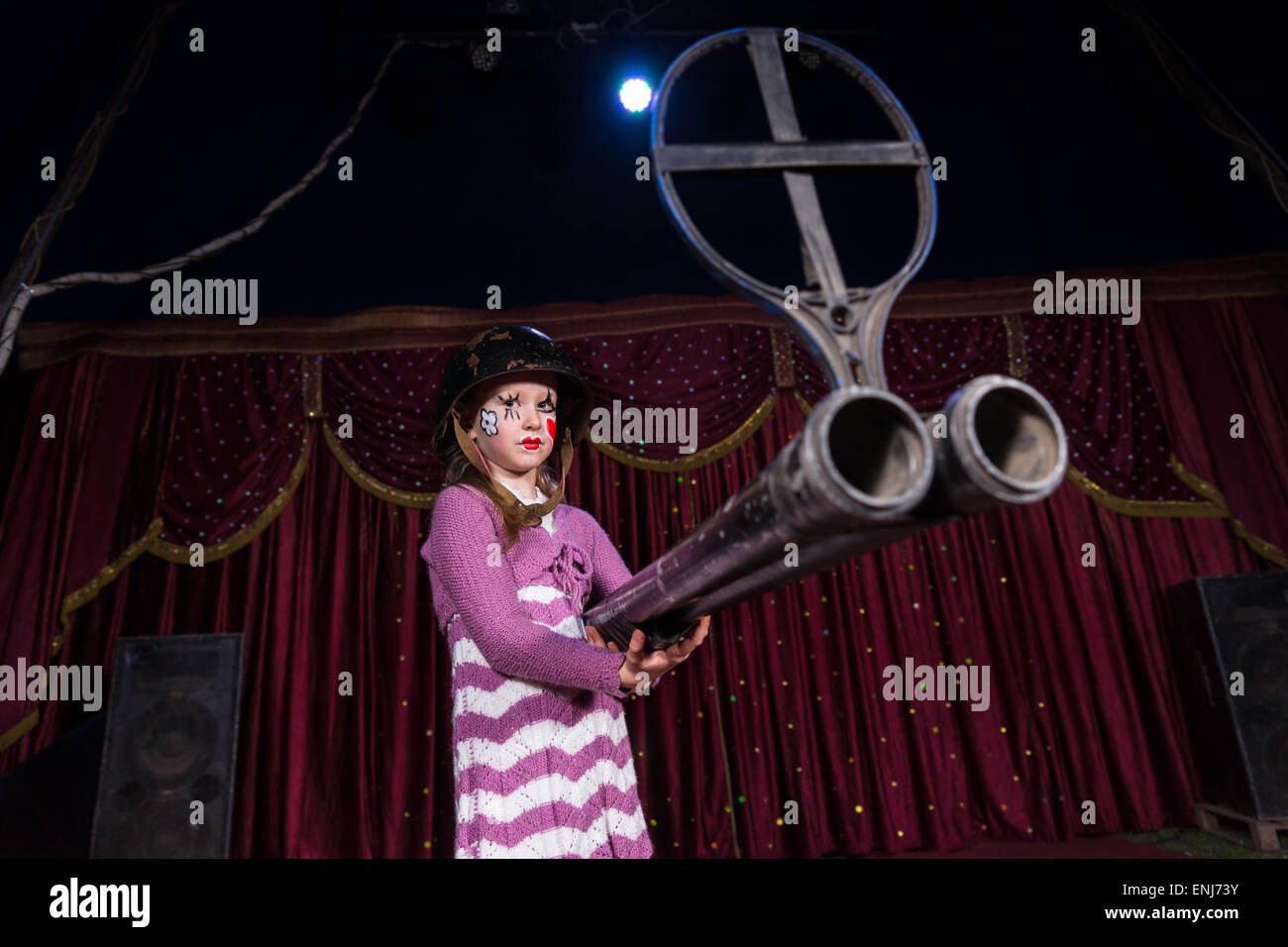 Girl with Painted Face Wearing Over Sized Helmet Holding Large Double Barreled Gun on Stage with Red Curtain Stock Photo