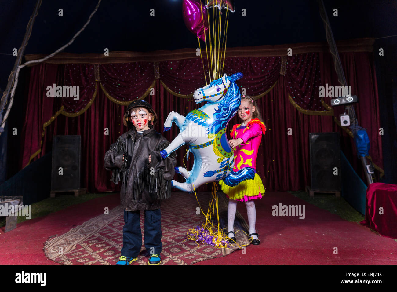 Boy Dressed as Clown Wearing Leather Jacket and Helmet Standing on Stage with Girl Wearing Bright Costume Holding Balloon String Stock Photo