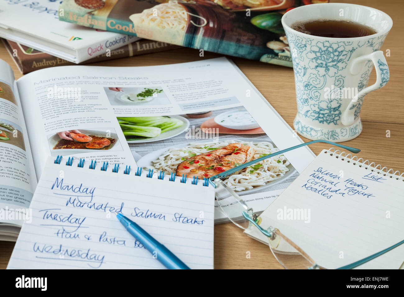 A notepad with a healthy eating meal plan for the coming week and a shopping list on a tabletop beside an open cookery book and a mug of black coffee. Stock Photo