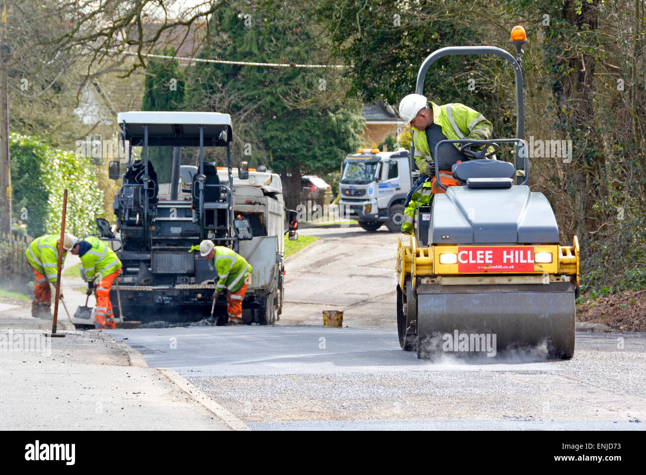 Men working at road works resurfacing & repairs using vibrating roller & tarmac spreading machine wearing high visibility jackets & hard hats Essex UK Stock Photo