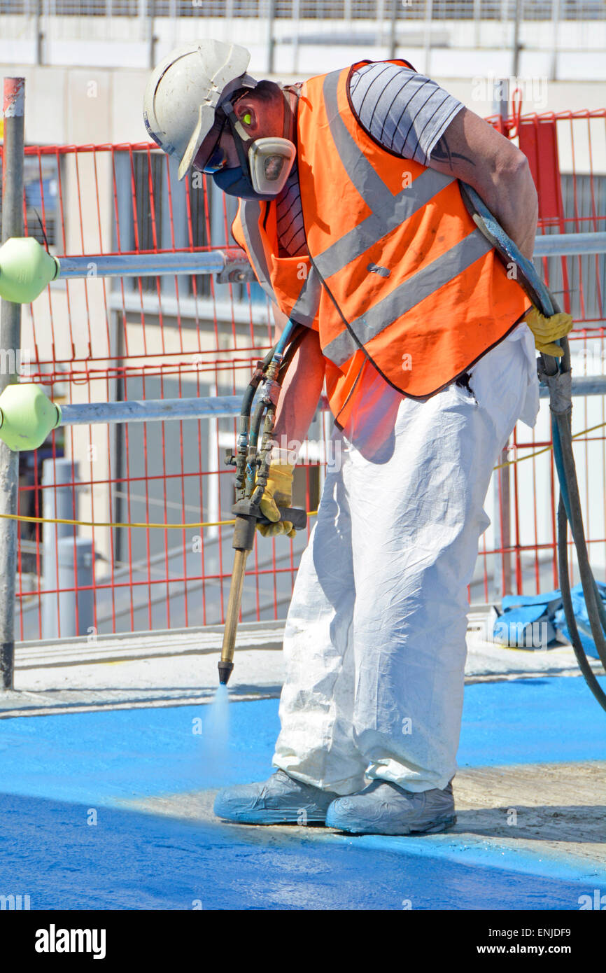Close up of worker protected by face mask & safety gear pressure spray painting blue coating on concrete floor high vis jacket & hard hat  London UK Stock Photo