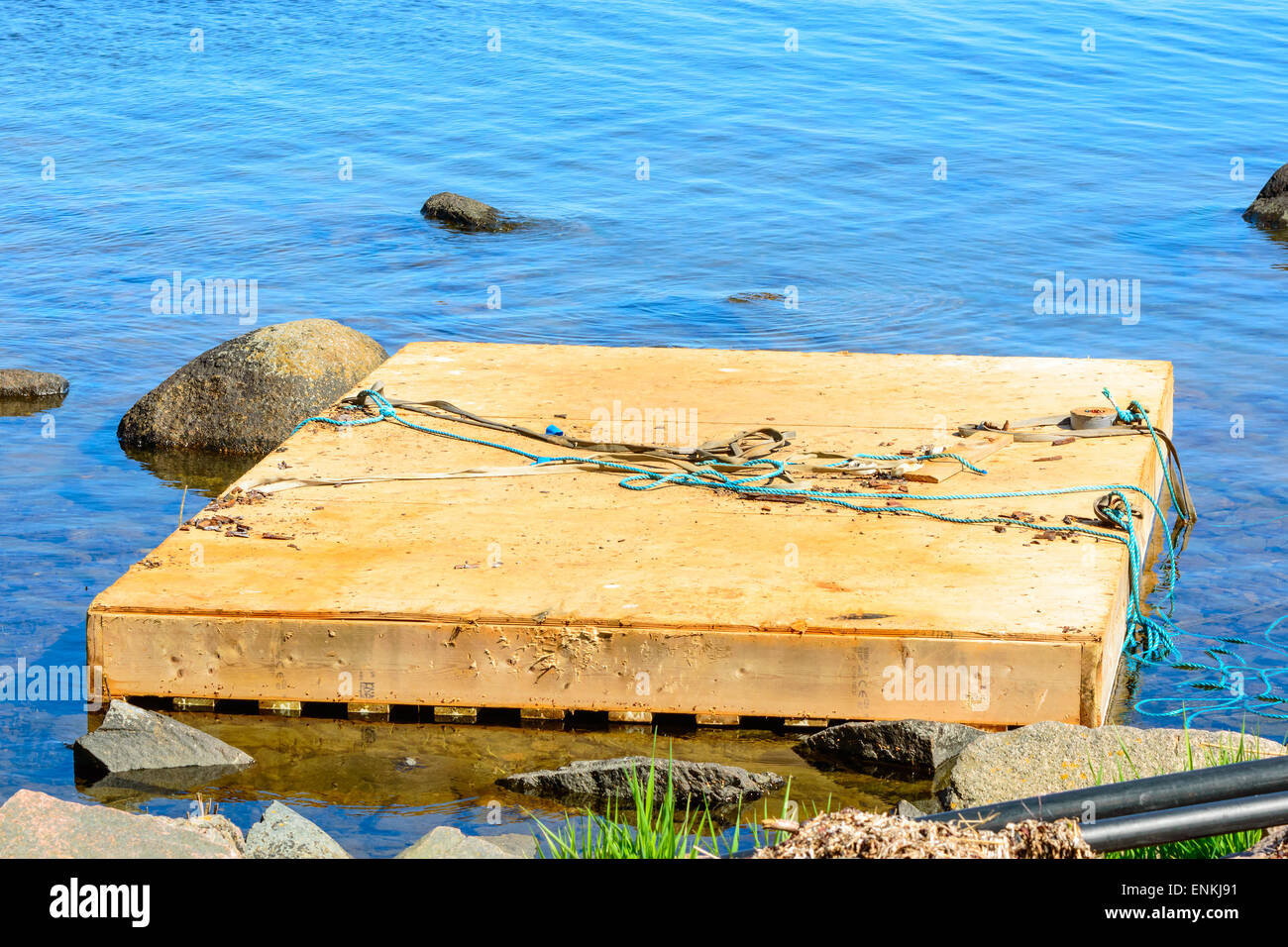 Wooden floating work platform by the shore close to stones. Blue water around and some ropes on the platform. Stock Photo