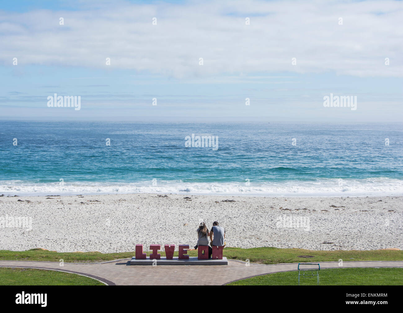 A couple at Camps Bay beach sitting on concrete letters spelling live on Stock Photo