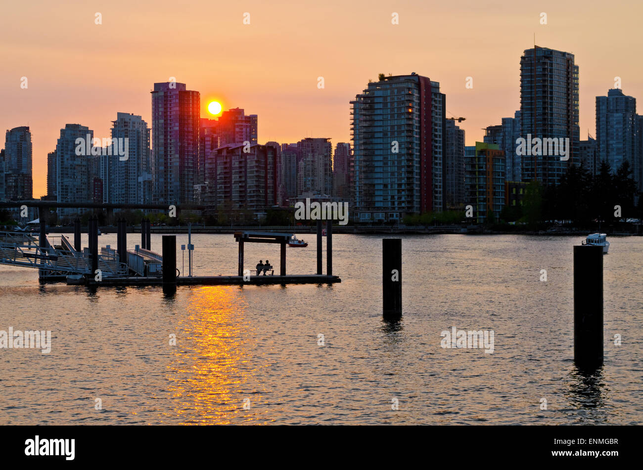 Beautiful sunset on False Creek in Vancouver, with the city buildings in silhouettes.  Two people sitting on a bench on the pier Stock Photo