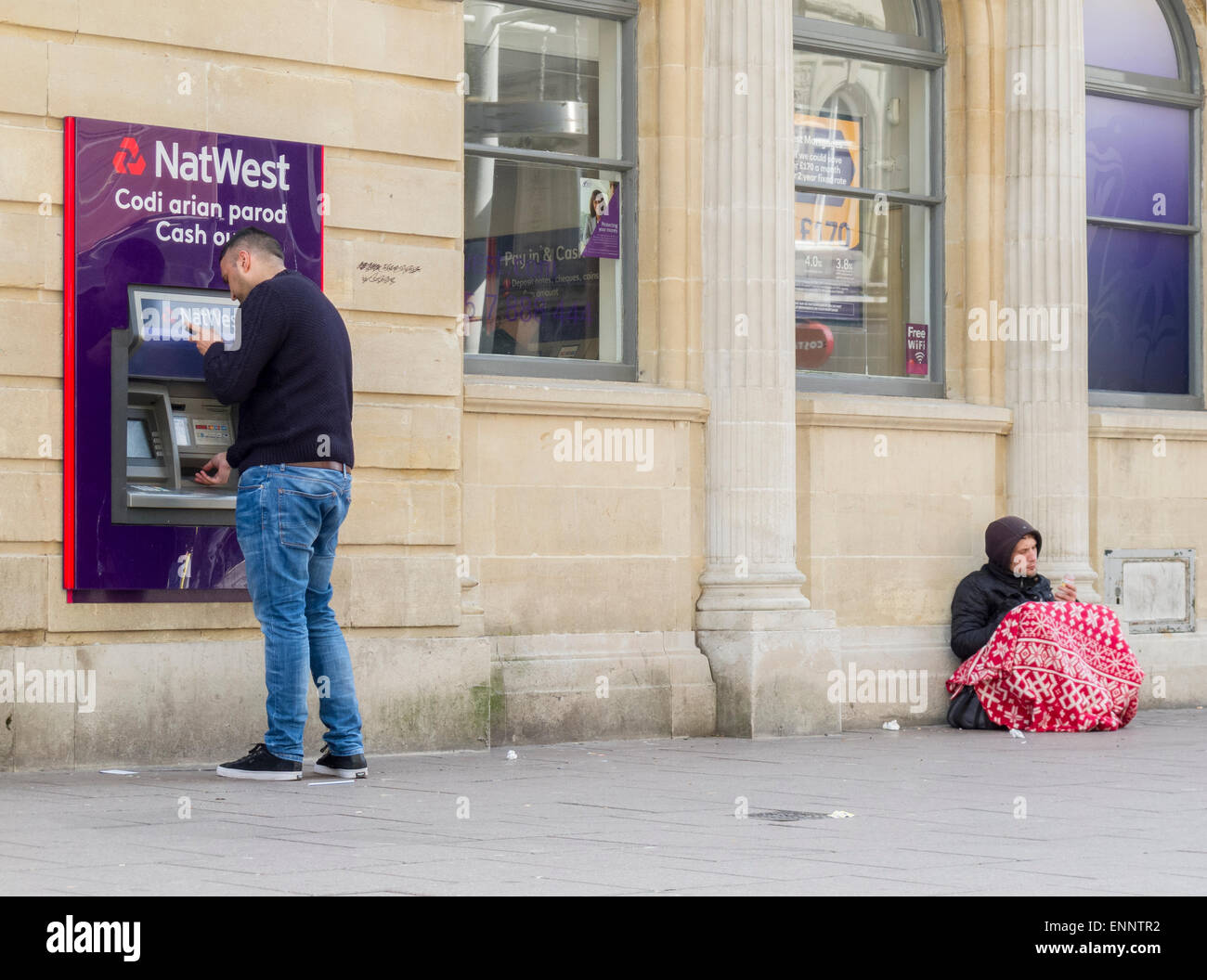 Beggar seated near a teller machine where cash is being withdrawn Stock Photo