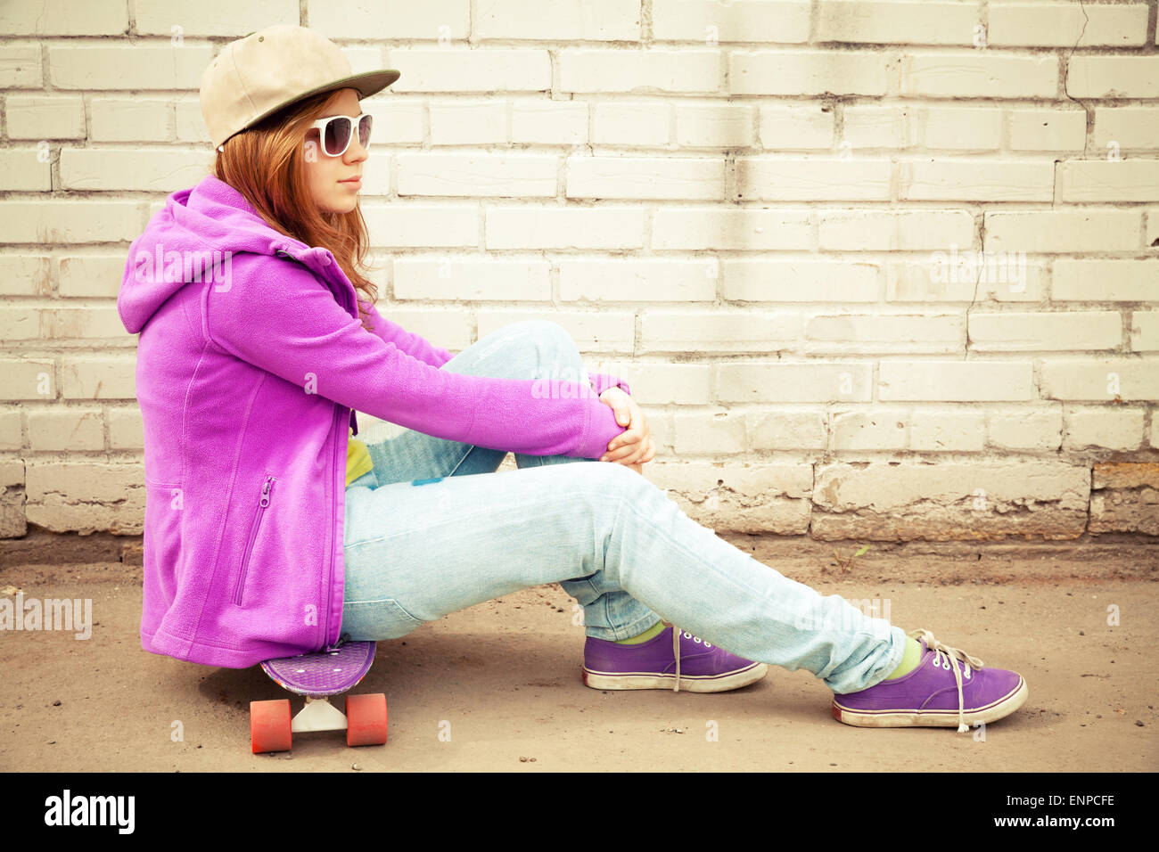 Blond teenage girl in jeans, cap and sunglasses sits on her skateboard near urban brick wall Stock Photo