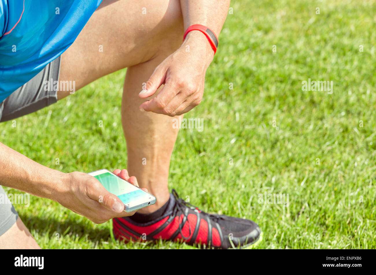 Sportive man kneels on a lawn and checks his fitness results on a smartphone. He wears a fitness tracker wristband on his left Stock Photo