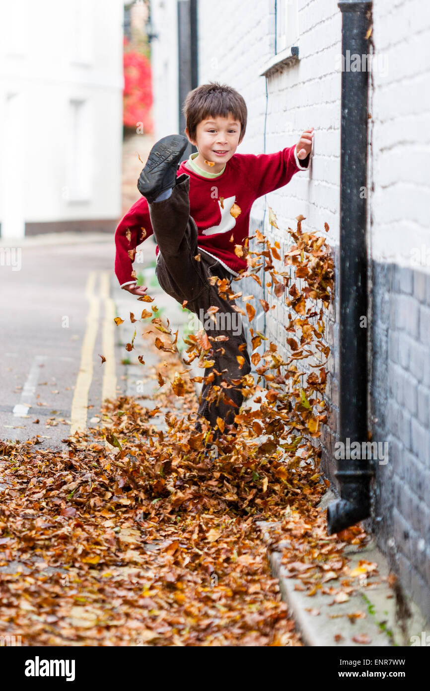 Caucasian child, boy, 8-9 year old, walking along a leave covered road towards the viewer, kicking the leaves high up into the air. Autumn. Stock Photo