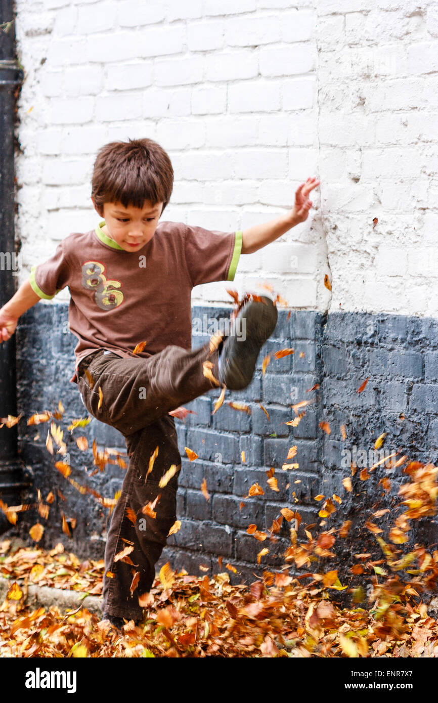 Caucasian child, boy, 8-9 year old, walking along a leave covered road towards the viewer, kicking the leaves high up into the air. Autumn. Stock Photo