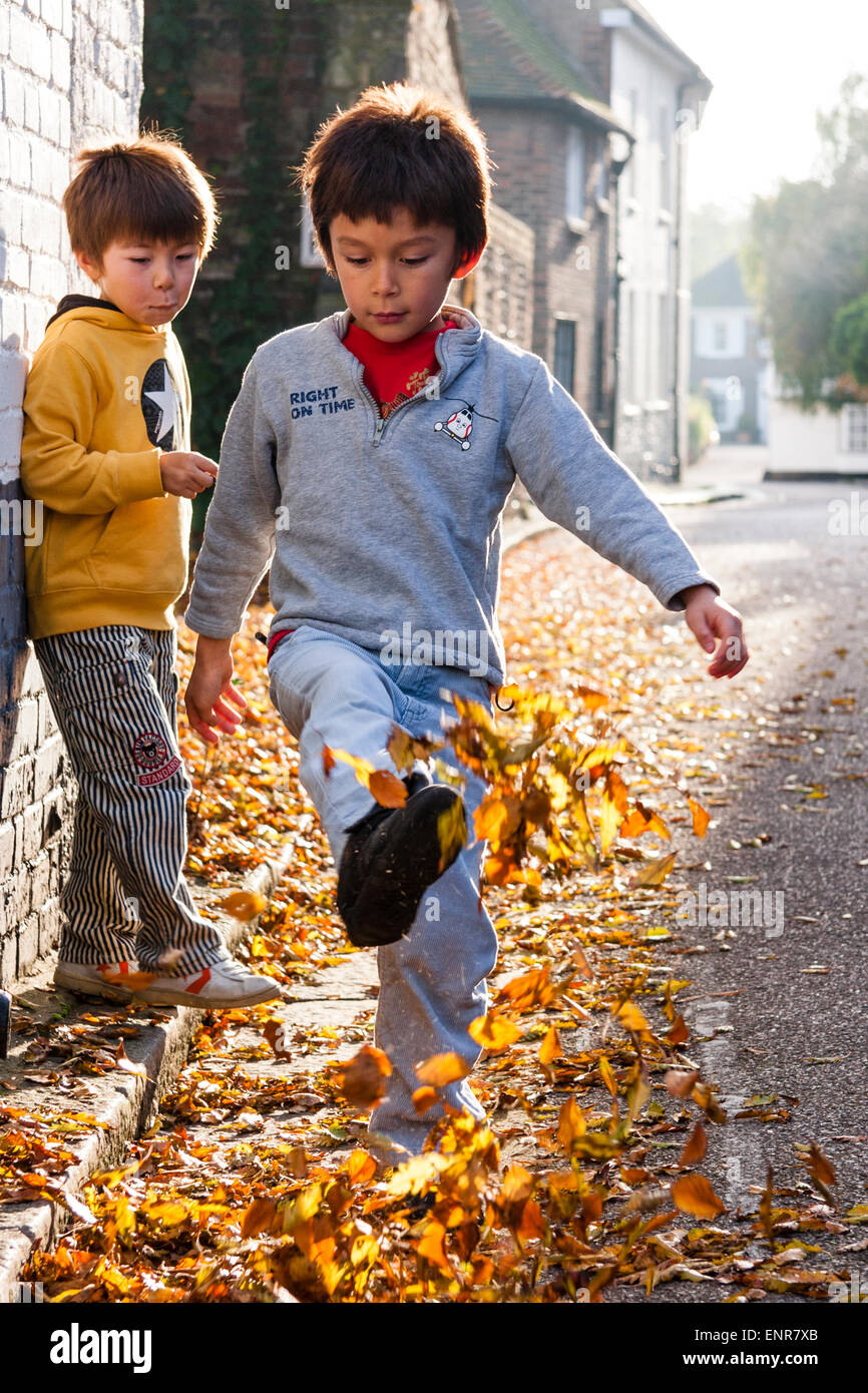 Caucasian child, boy, 7-8 year old, walking along a leave covered road towards the viewer, kicking the leaves into the air while another boy watches. Stock Photo