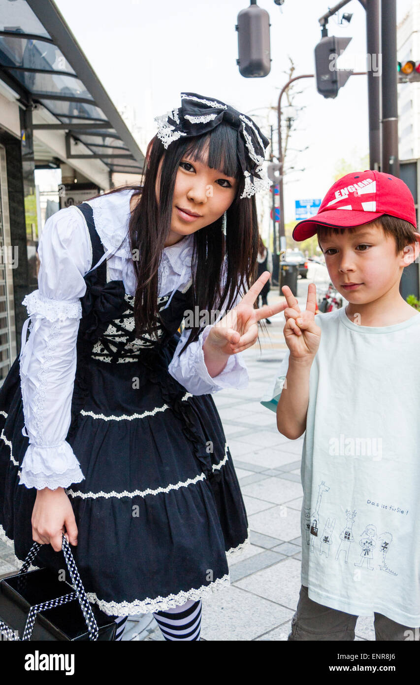 Japan, Kyoto. Costume play girl, Goth Lolita Victorian Maid black outfit. Posing with Asian-Western mixed race child. Both giving peace gesture. Stock Photo